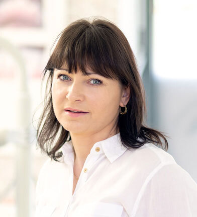 Frau mit braunen Haaren und weißer Bluse blickt freundlich in heller Praxisumgebung.