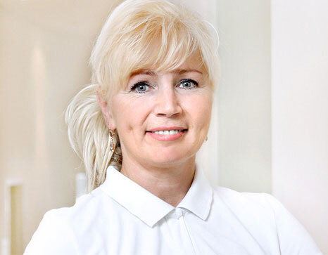 Porträt einer lächelnden Frau mit blonden Haaren in weißer Bluse in heller Praxisumgebung.