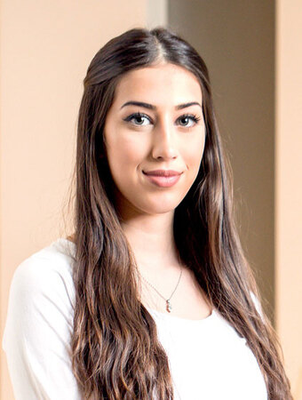 Junge Frau mit langen braunen Haaren lächelt freundlich in einem hellen Innenraum.